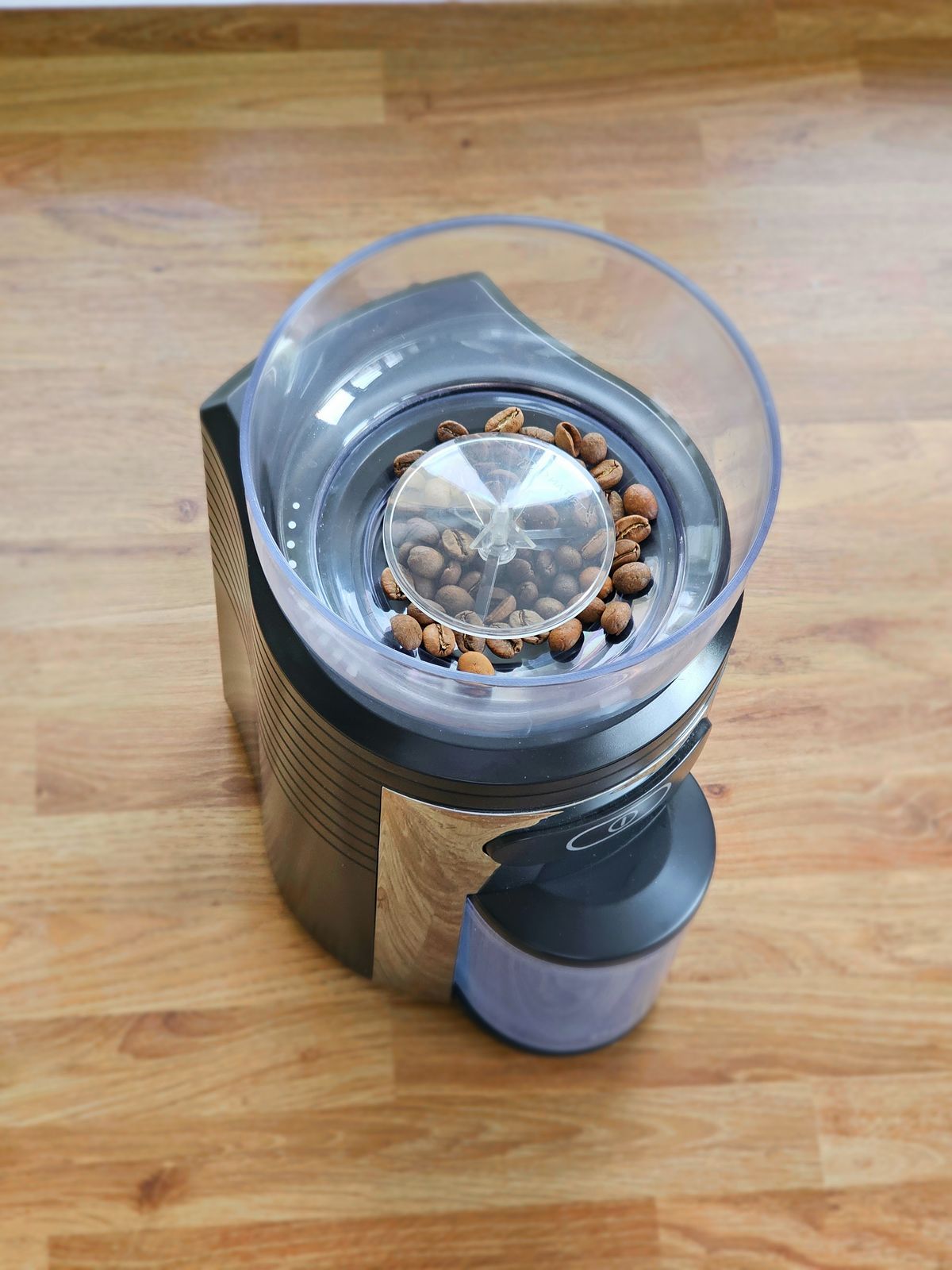 A Dualit CCG2 conical burr grinder on a wooden floor, seen from above, with roasted beans in its clear hopper and the grounds container clipped into the front