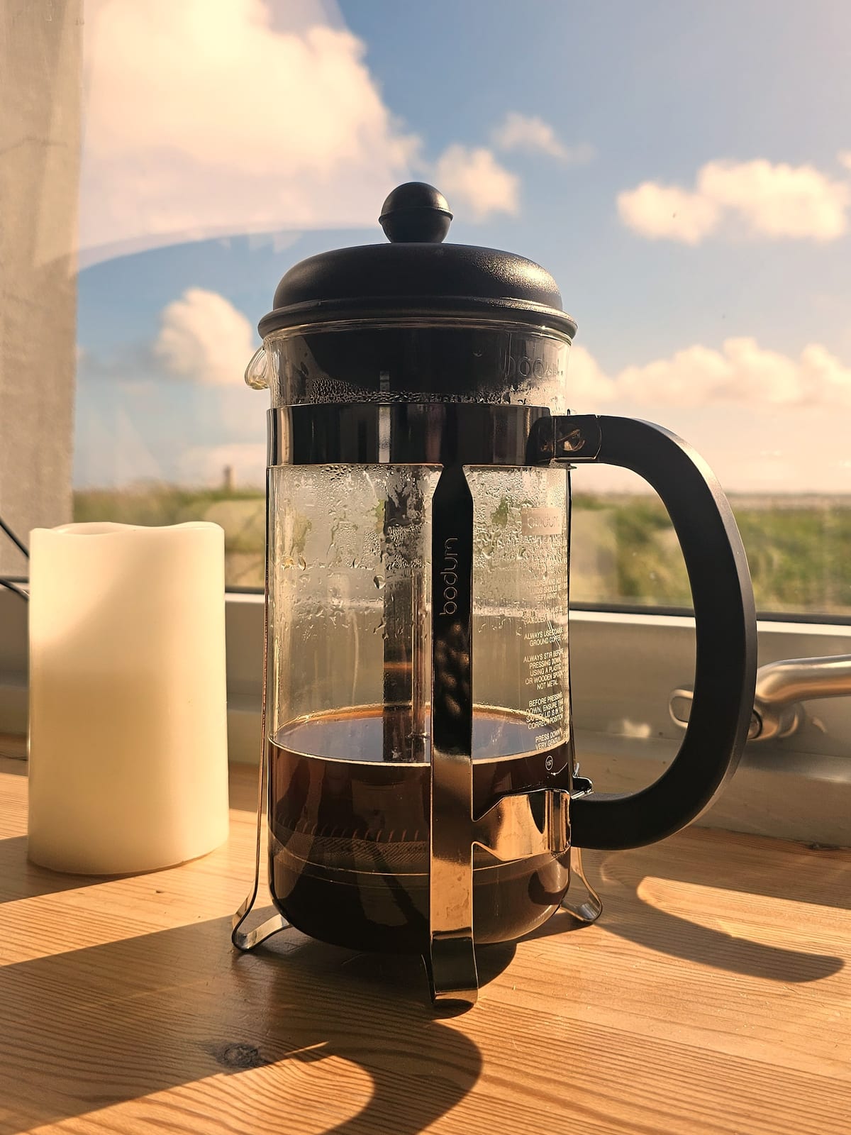 A Bodum cafeti&egrave;re of brewed coffee, plunger down, on a sunlit wooden table beside a candle, with a bright sky through the window behind