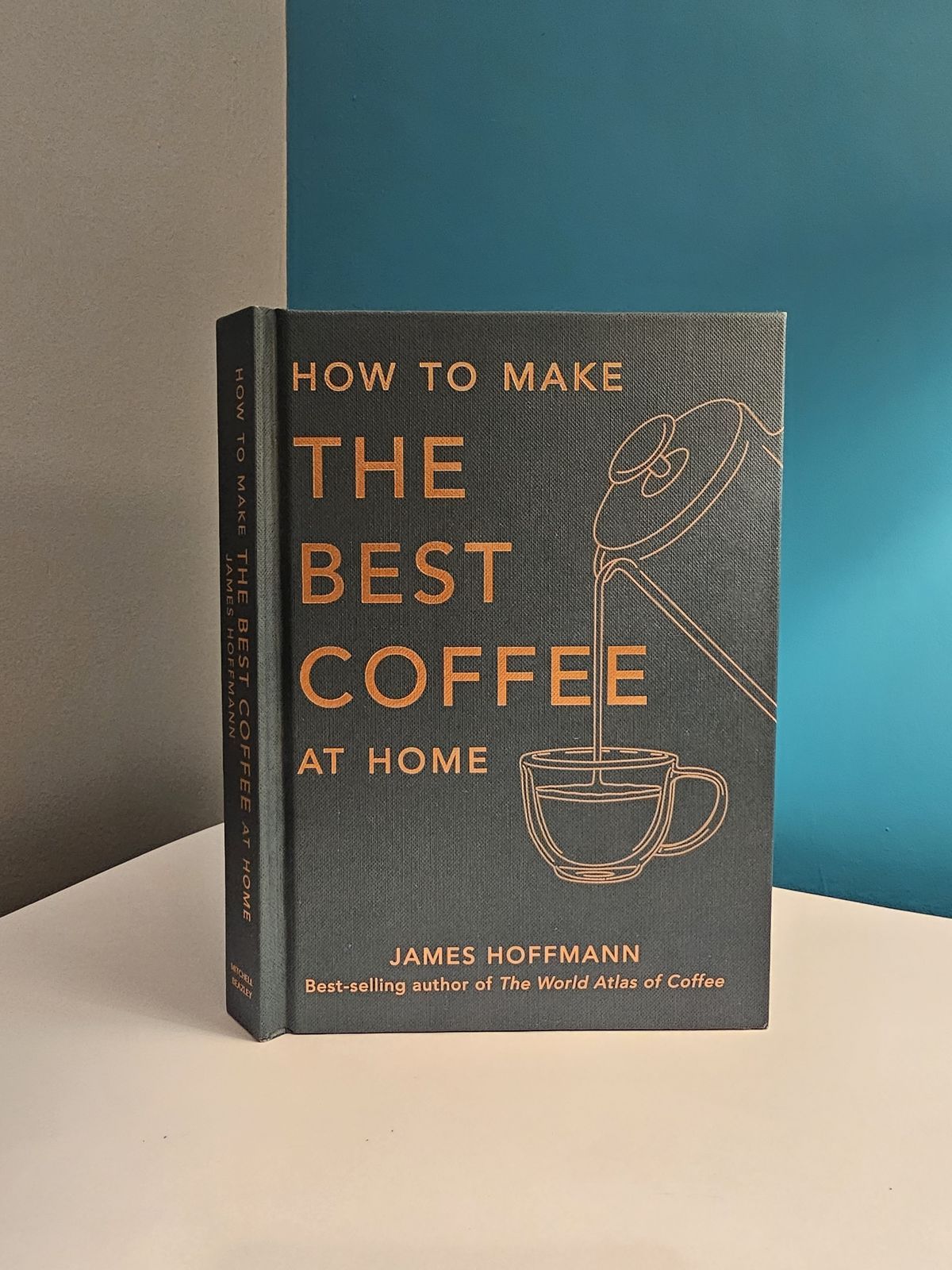 A hardback copy of James Hoffmann's How to Make the Best Coffee at Home standing on a white surface in the corner where a grey wall meets a teal one, its slate cover showing a line drawing of a cafeti&egrave;re pouring into a cup