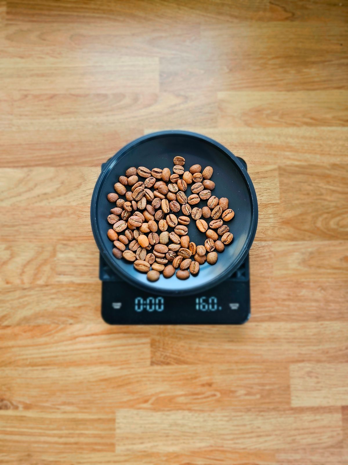 A black coffee scale seen from above on a wooden table, a dish of roasted beans on its platform and the display reading 0:00 on the timer and 16.0 grams