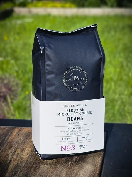 A black bag of M&S Collection single origin Peruvian micro lot coffee standing on a weathered wooden table with grass behind it
