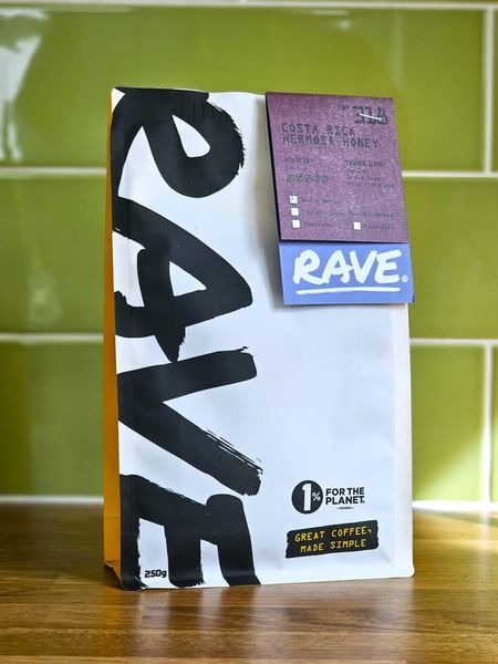 A 250g bag of Rave Coffee standing on a wooden worktop against green tiles, its label reading Costa Rica Hermosa Honey